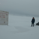 A man stands next to a small snowmobile on an icy landscape, staring at a box covering an oil test well.